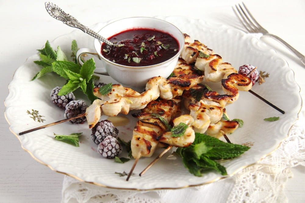 Skewers of grilled chicken on a plate with greens and a white bowl of dark purple blackberry sauce.