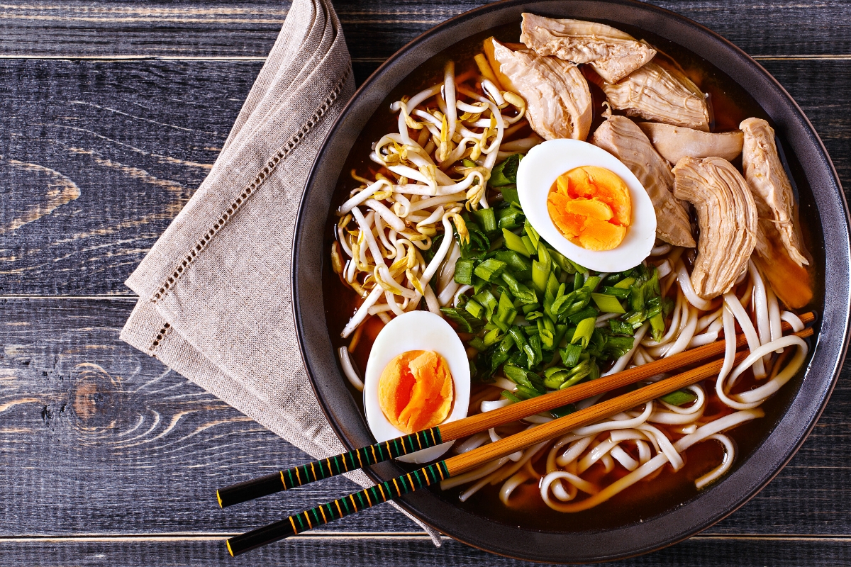 Bowl of ramen topped with sliced chicken, soft boiled egg halves and snipped chives with chopsticks resting on it.