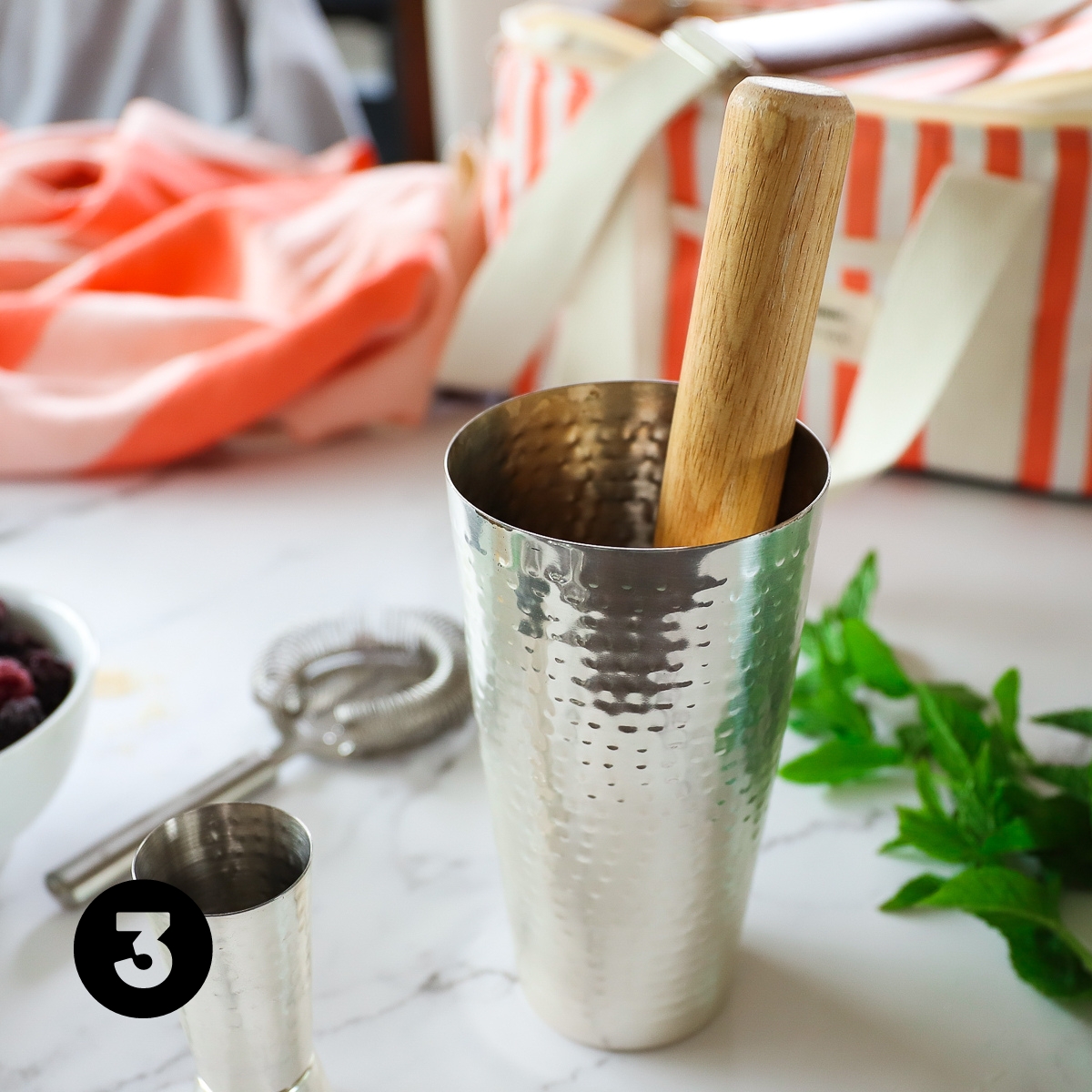 Side view of the hammered silver cocktail shaker with the wooden muddler in it. Mint leaves are beside it.