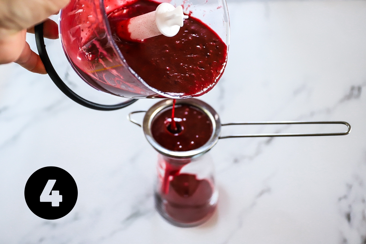 The deep red dressing is being strained through a metal sieve into a glass jar.