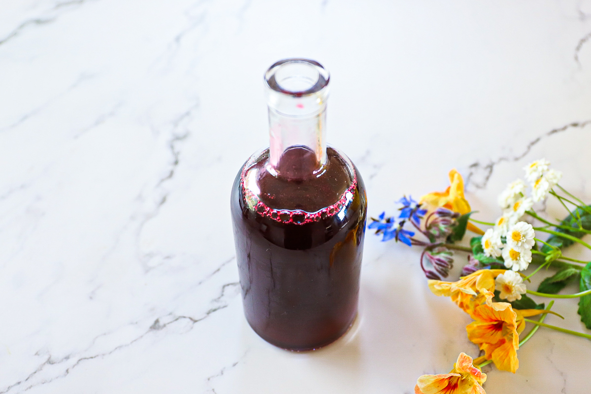 Glass bottle with a small neck full of dark purple blackberry syrup with flowers on a white table.