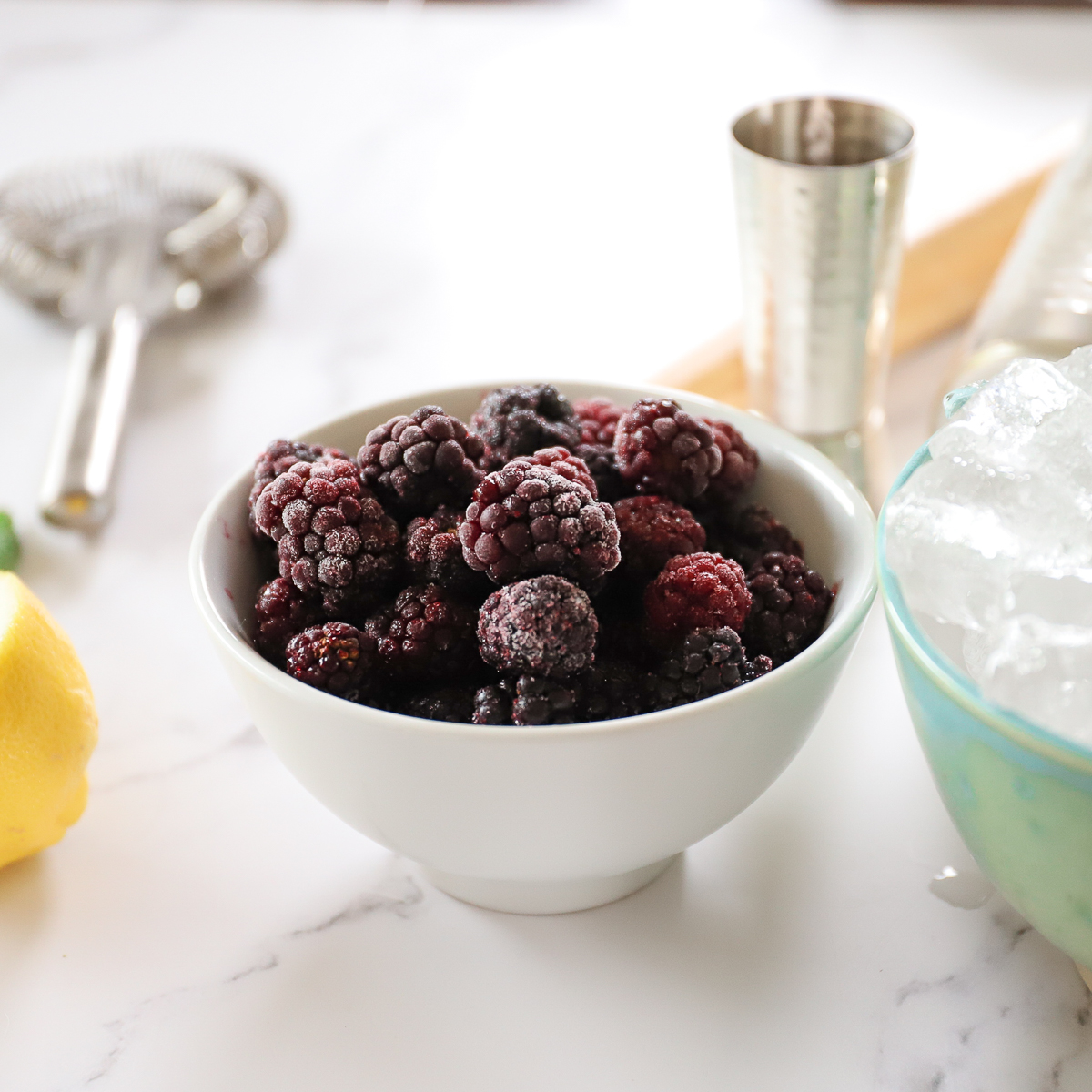A white bowl of frosty looking blackberries with ice cubes, a jigger and cocktail strainer on the table around them.