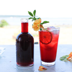 A bottle of dark purple juice beside a glass with a lighter purple-red drink with bubbles in it. It is garnished with a lemon wedge, mint sprig and pale orange nasturtium flower.