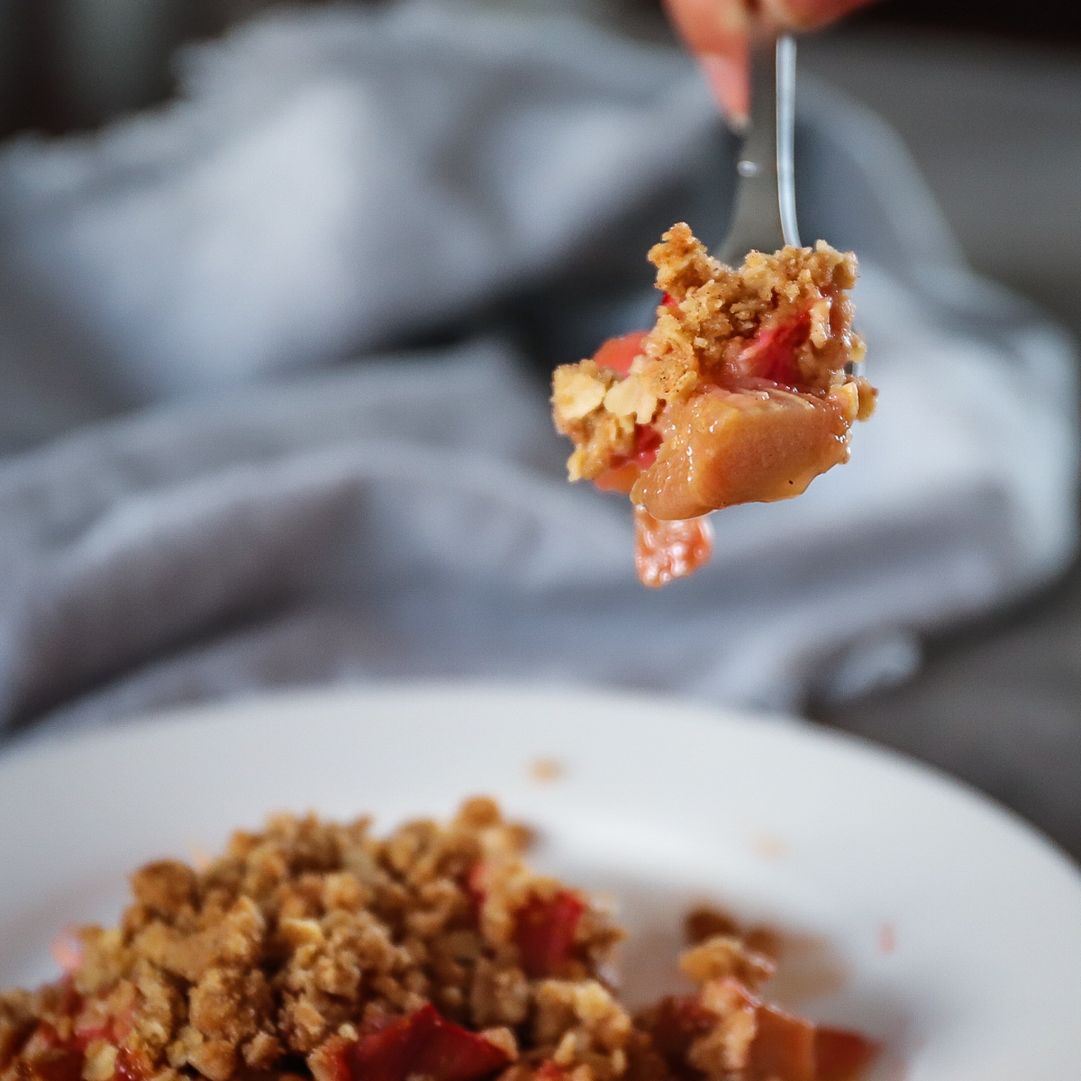 A forkful of rhubarb crumble being lifted up with juicy drips of rhubarb juice spilling off.