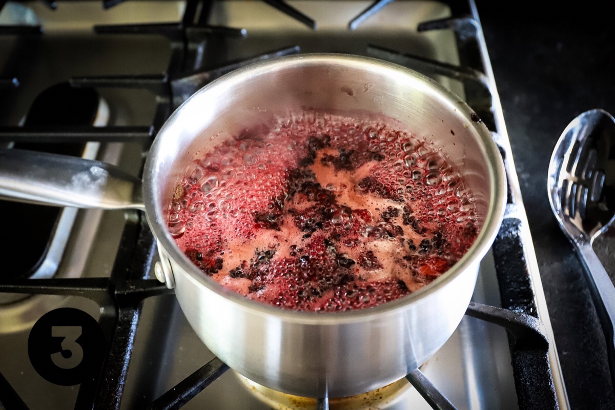 The pot of berries and sugar in the pot are at a medium boil with frothy, light pink bubbles on the top.