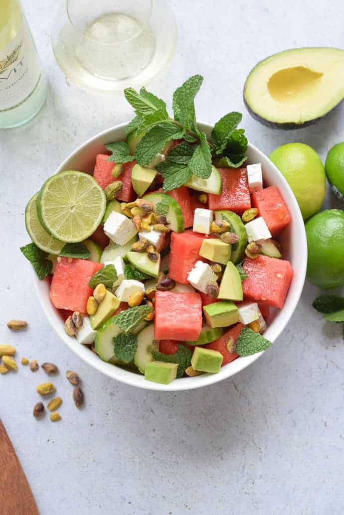 A white bowl of cubed watermelon, feta cheese and cucumbers with some lime wheels on the edge.