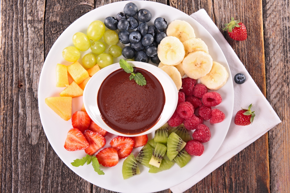 Plate of blueberries, banana slices, raspberries, chopped kiwi, strawberries, cubed cantaloupe and grapes around a small dish of chocolate dip.