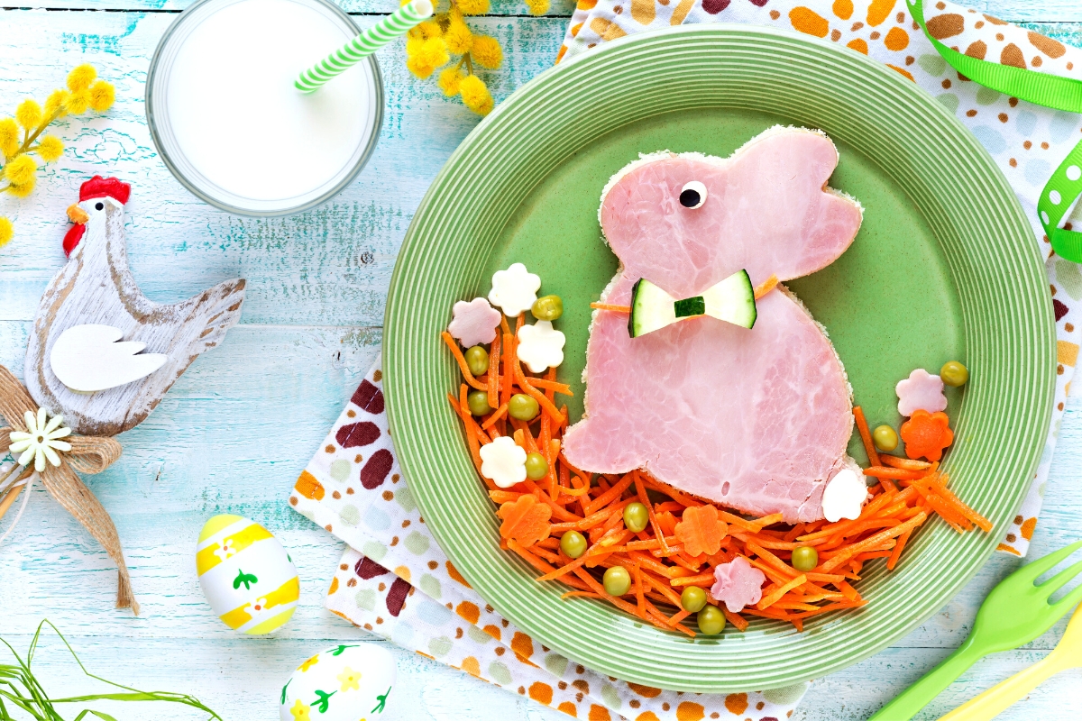 A open faced ham sandwich in the shape of a bunny rabbit with shredded carrot grass under it on a green plate.