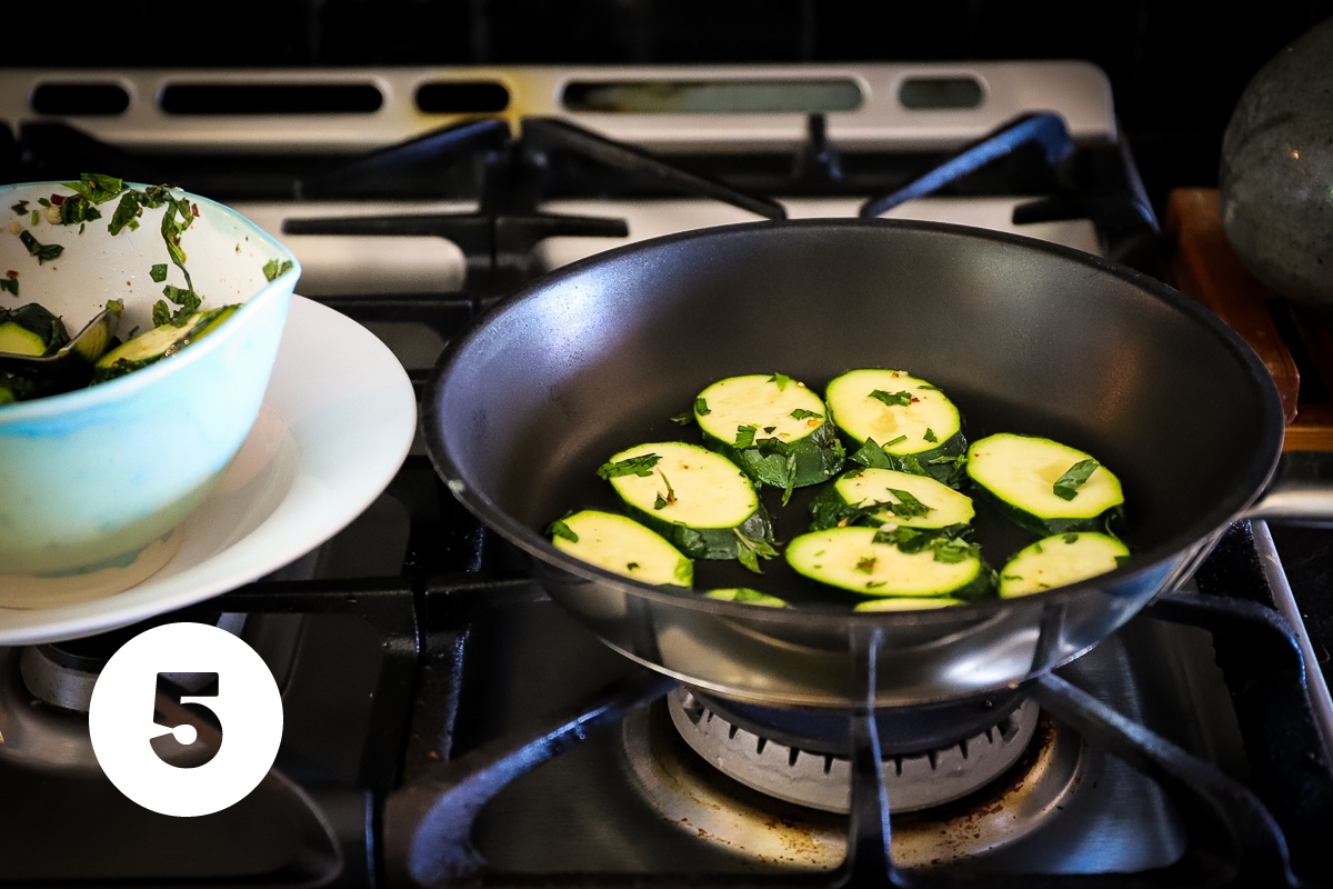 A black pan on a gas stove has raw zucchini slices in a tight layer on the bottom.
