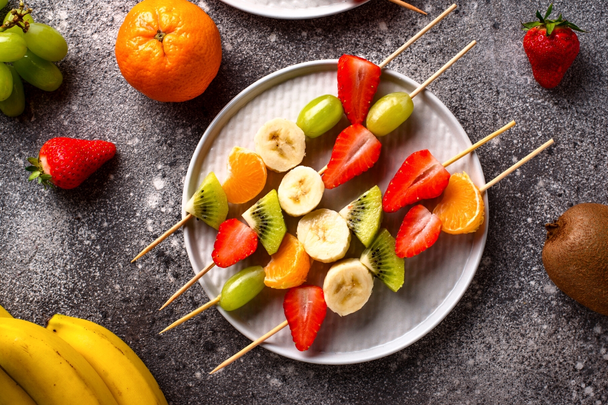 Wooden skewers with strawberries, banana, green grapes and orange segments threaded on and served on a round white plate.