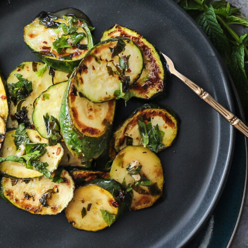 Pan fried oval slices of green zucchini on a black plate with a scattering of green herbs.