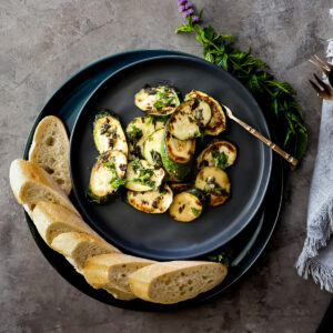 Marinated and fried zucchini slices with brown pan marks and green herbs on a black plate with sliced bread around one side.