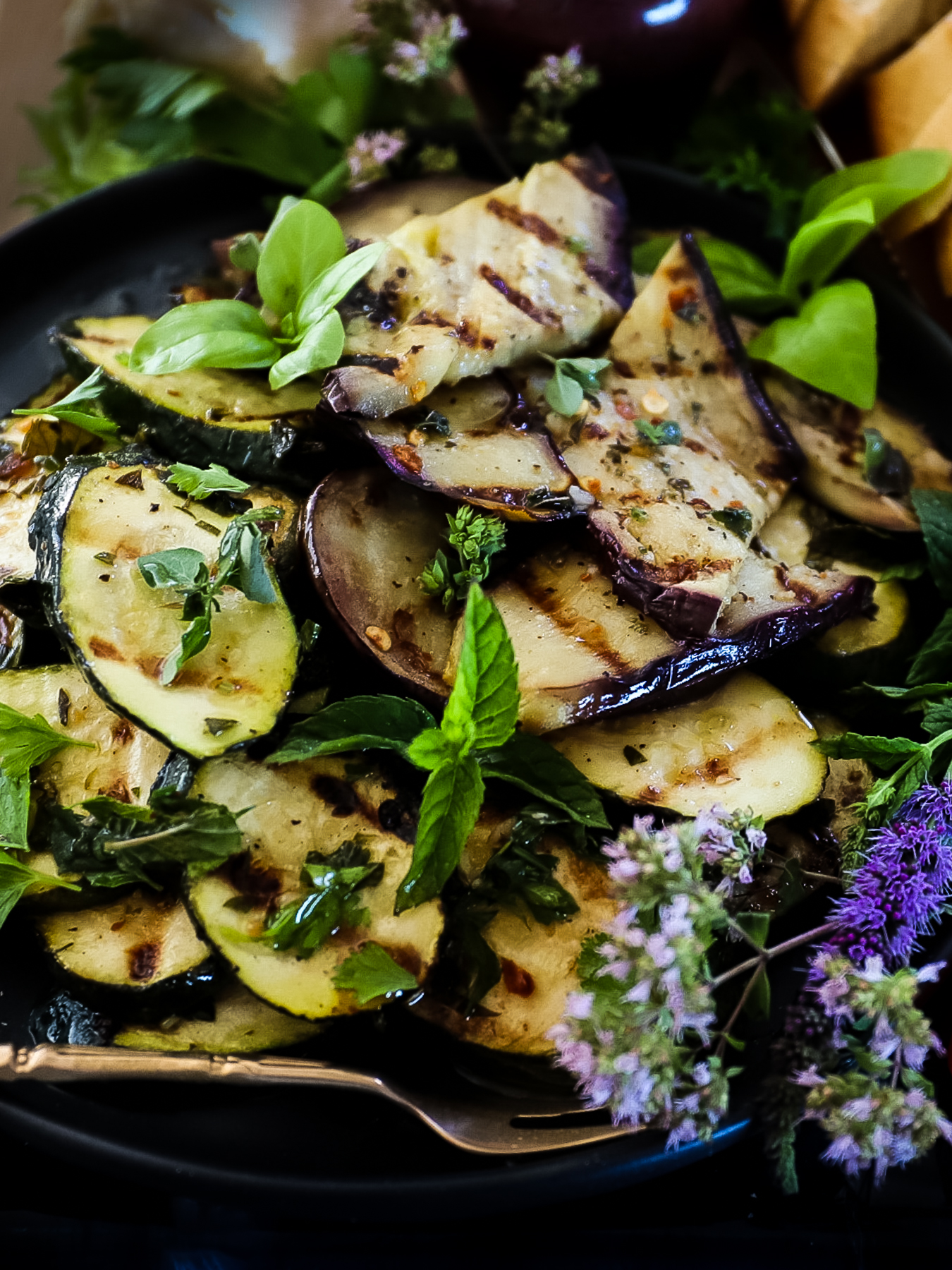 Close up of grilled zucchini and eggplant with grill marks on each and flowering oregano and mint leaves garnishing it.