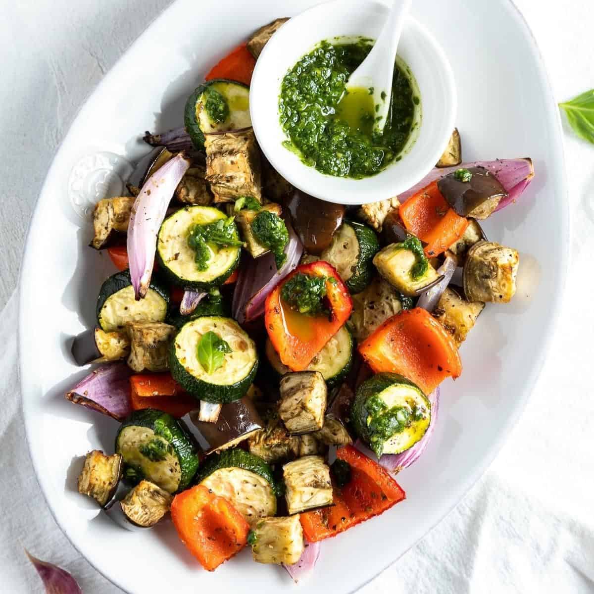 White oval serving dish of colourful roasted vegetables with a small dish of green herb sauce on the side.