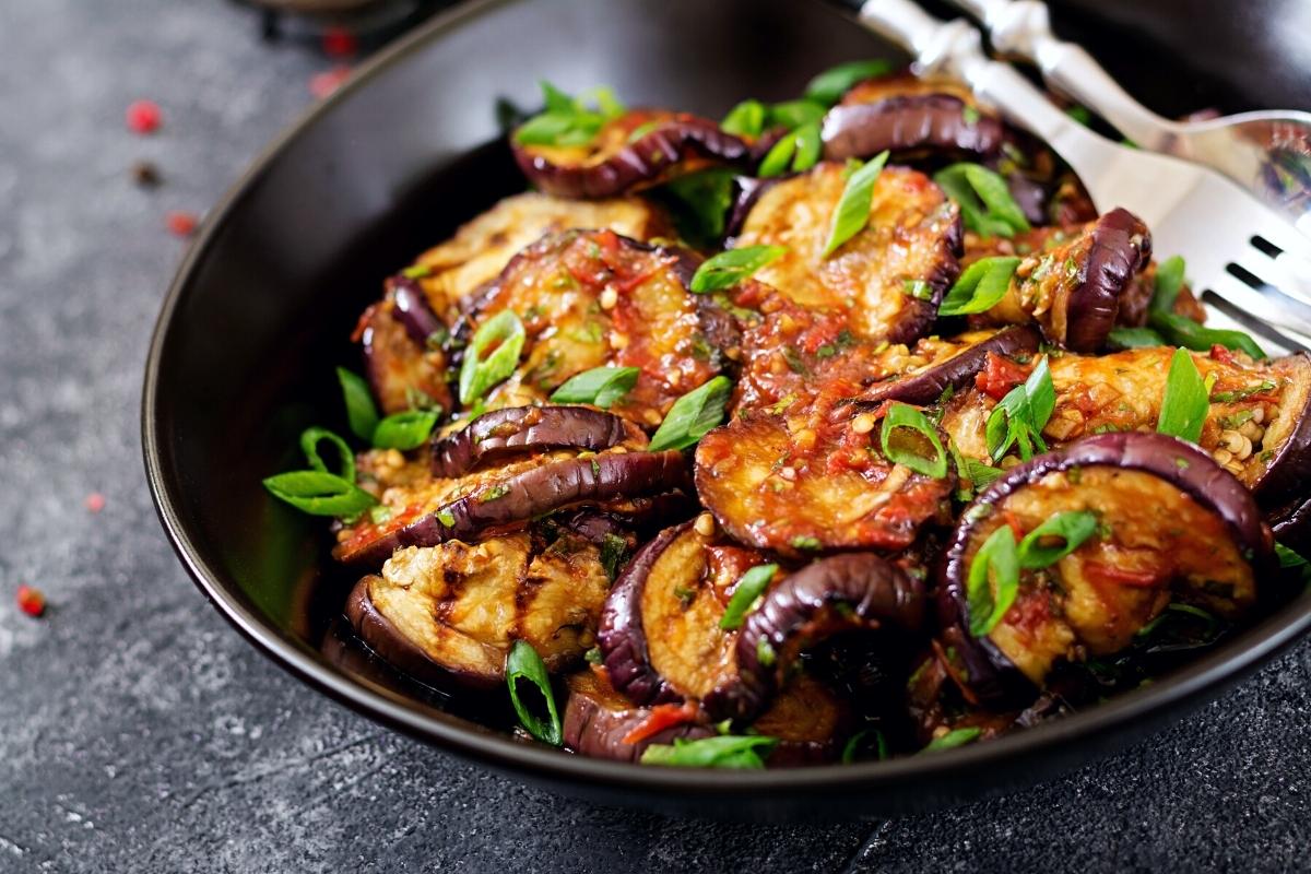 Sautéed eggplant in a black pan with tomato sauce and green herbs.