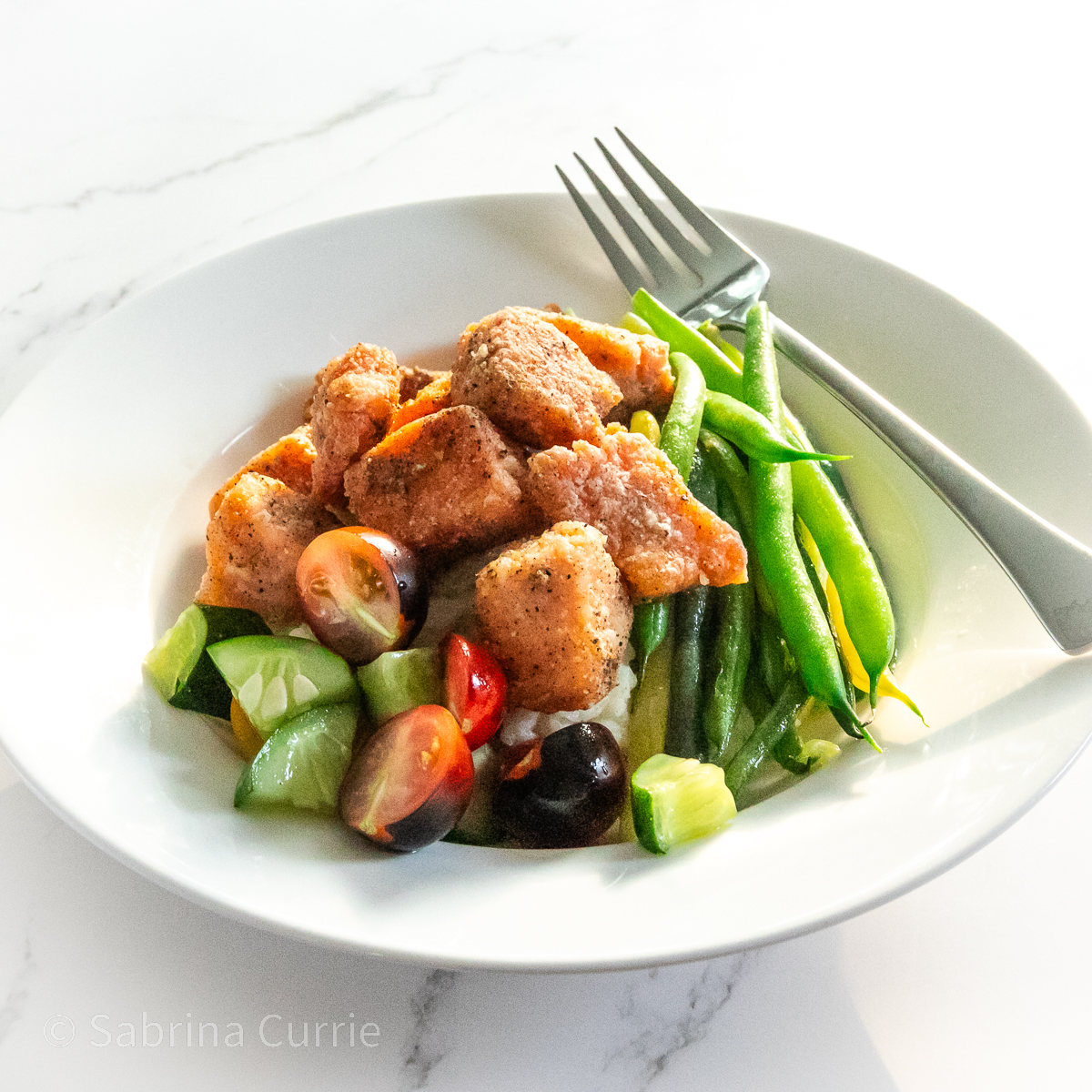 White, wide rimmed bowl with cubes of crispy salmon on top of a cucumber tomato salad on one side and green beans on the other side.