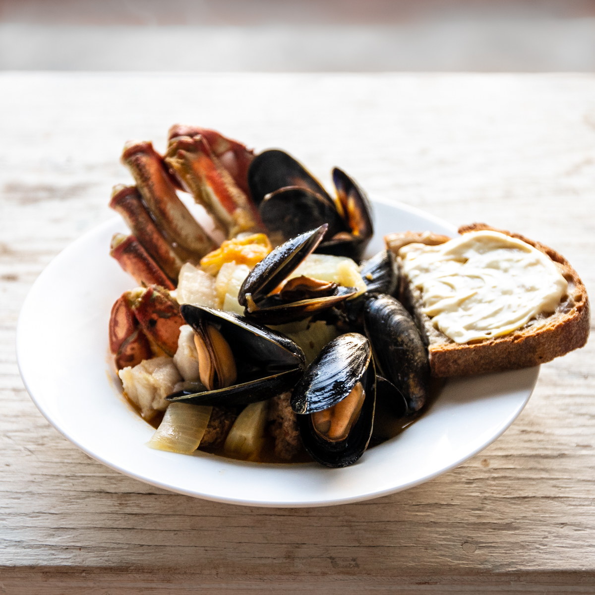 White, wide-rimmed bowl filled with crab, mussels and fish with a piece of aioli topped toast on the side.