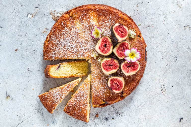 A round cake with 3 small wedges cut and sitting near is decorated with fresh cut figs on one quarter of the uncut cake.