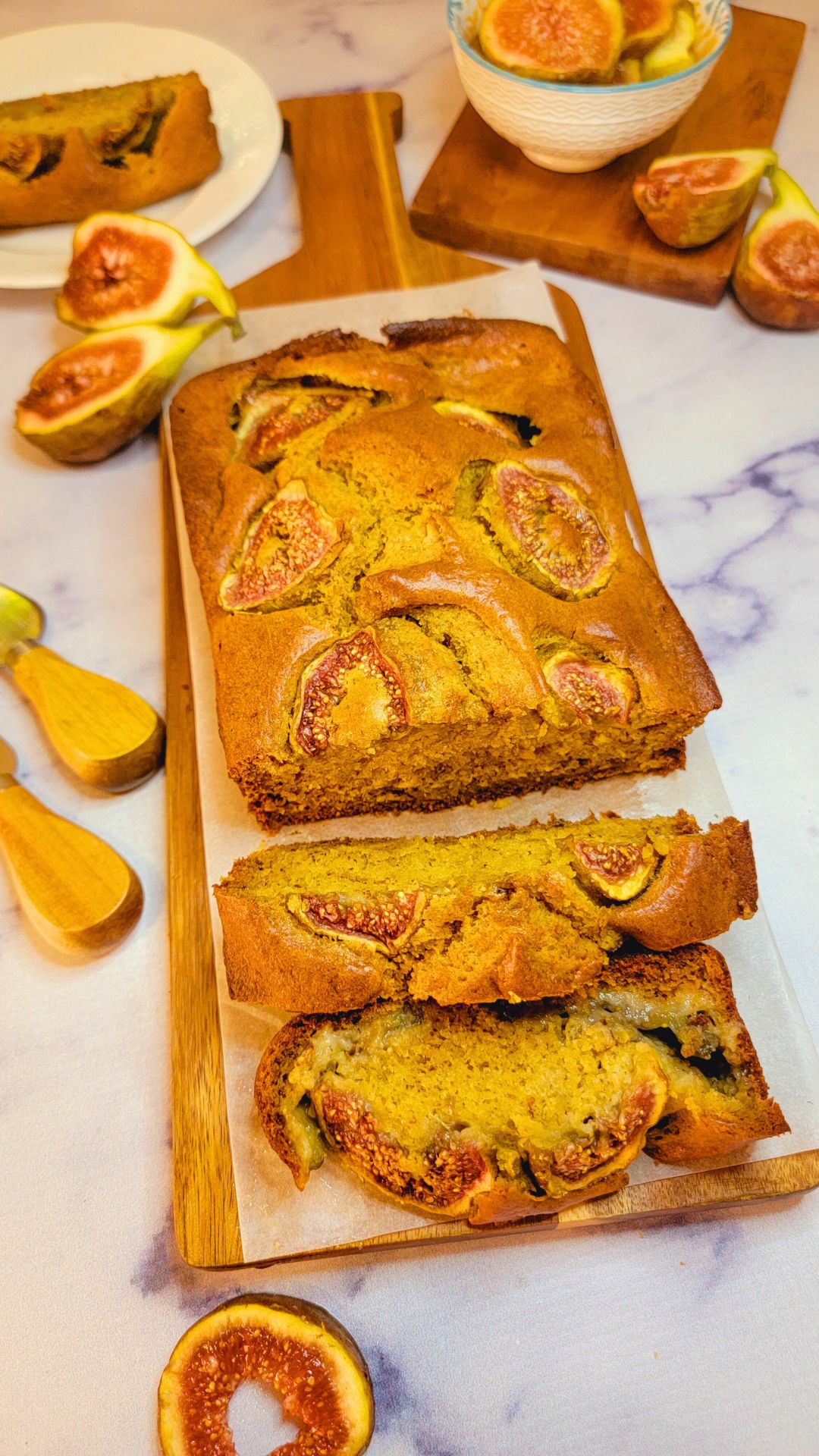 A loaf of fig bread with figs baked onto the top. Half the loaf is sliced with slices laying sideways on the cutting board.