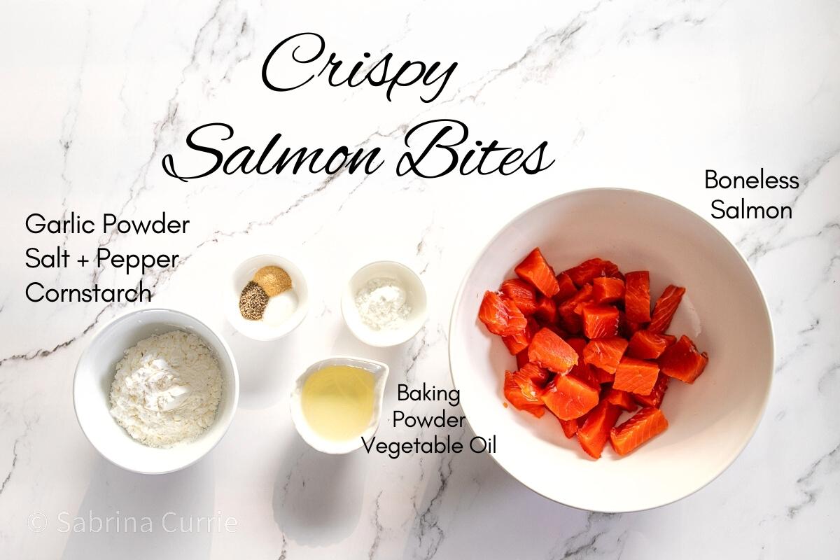 Ingredients for recipe with text labels. Ingredients include a white bowl of cubed red salmon, white cornstarch, pale yellow oil, baking powder and a small dish with salt, pepper and tan colored garlic powder.