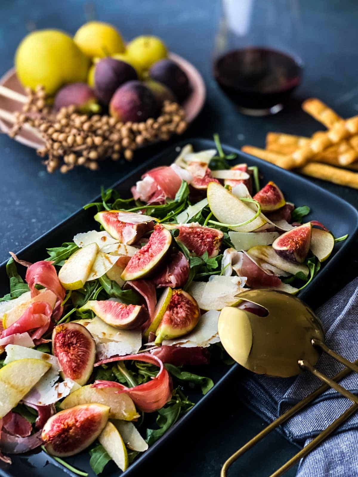 Sliced pears and figs on a dark platter with greens and a gold colored serving spoon.