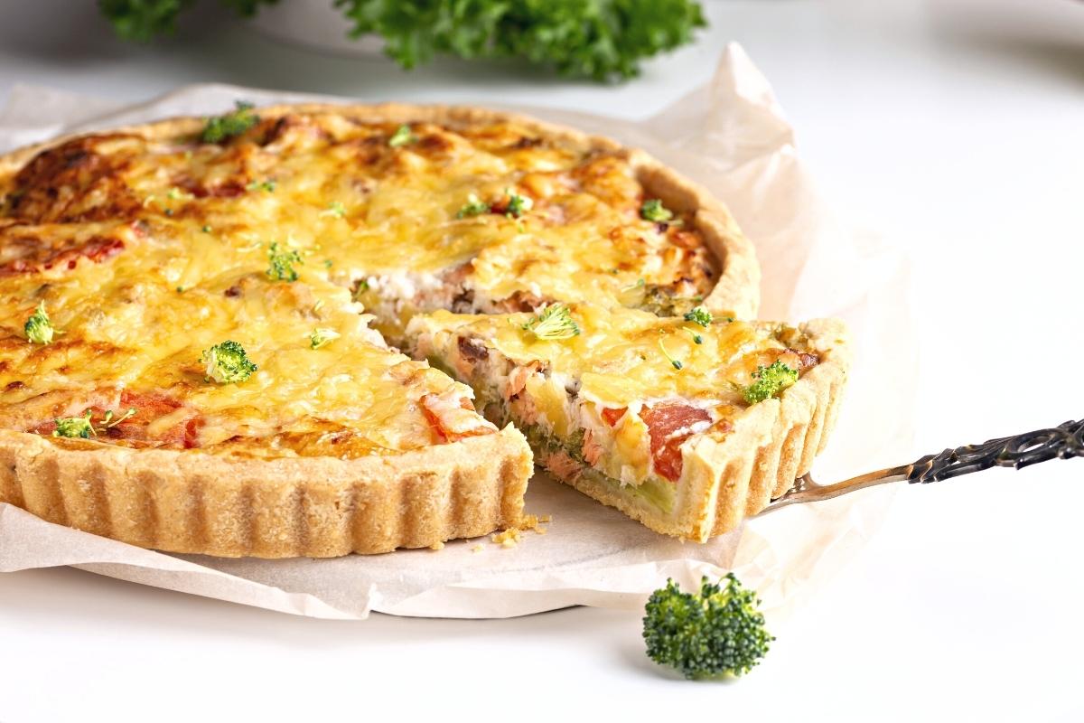 Salmon quiche with a slightly browned cheesy top and chunks of pink salmon inside. There is a small sprinkling of broccoli florets around.