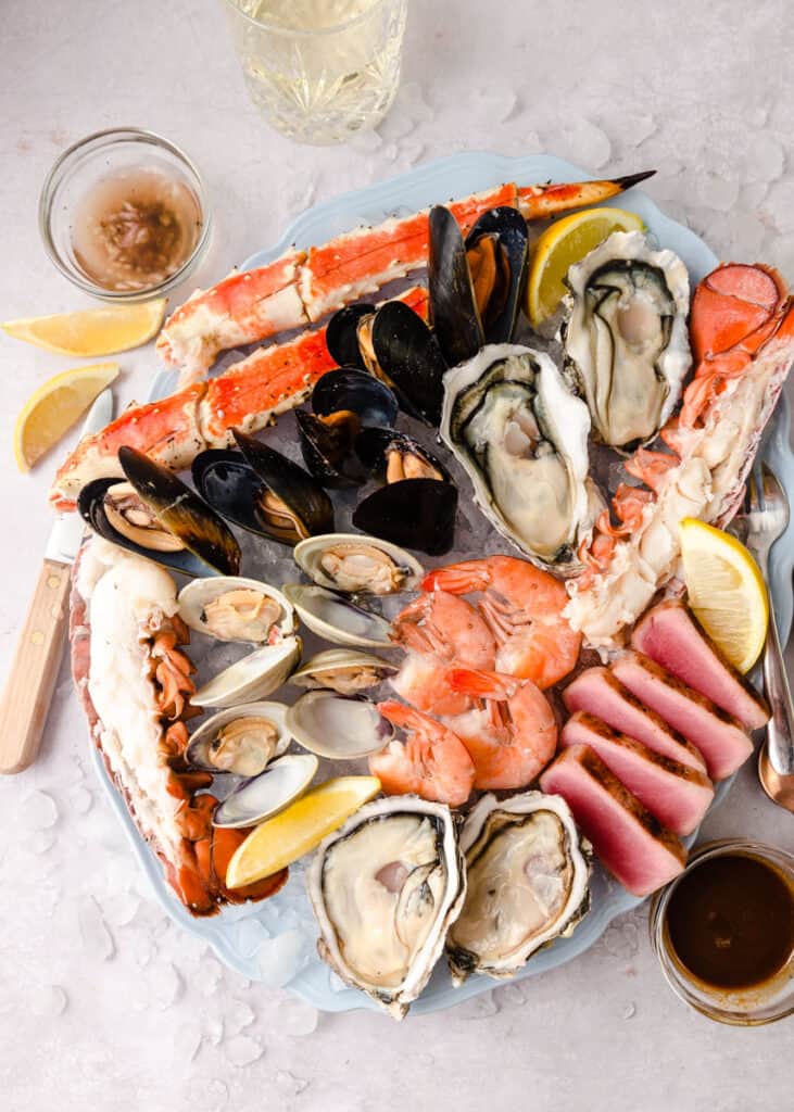 Round platter filled with assorted cooked seafoods including crab legs, shrimp, raw oysters on the half shell, thinly sliced raw tuna, clams in their shell and mussels.