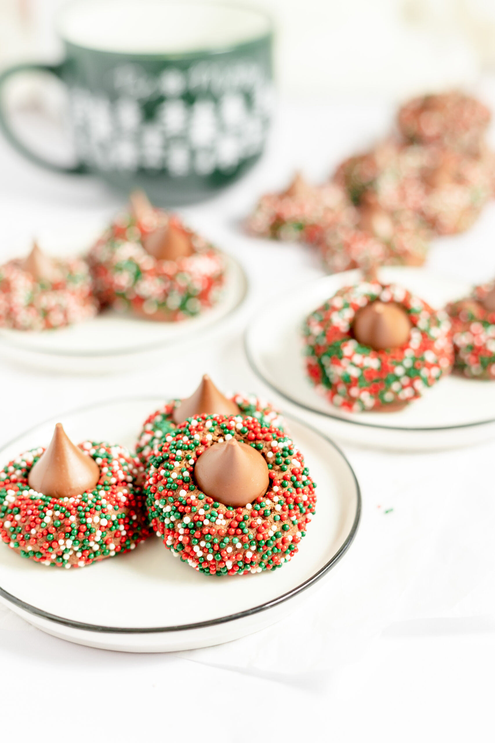 Four white plates with 3 chocolate cookies on each plate. The cookies are small, round and dipped in red, white and green sprinkles with a chocolate Hershey's kiss on top.
