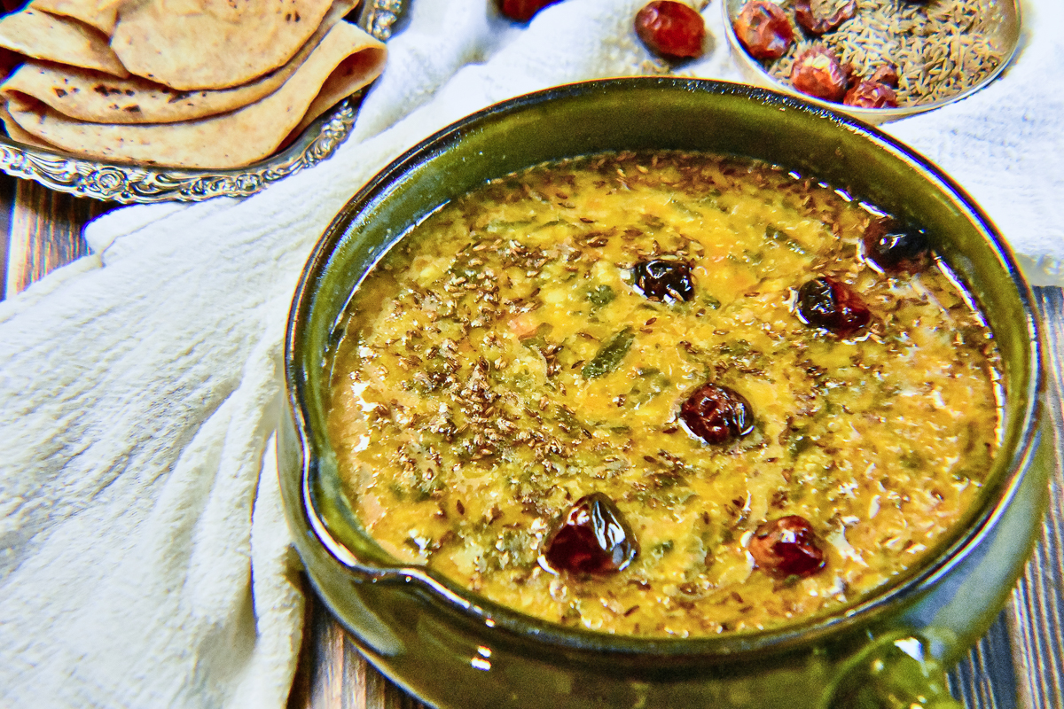 Looking into a dark green pot on a white tablecloth filled with with yellow-orange palak dal topped with cumin seeds and red button chilis.