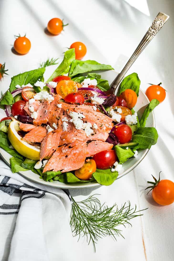 Chopped salad of green lettuce, red and yellow cherry tomatoes and large flaked salmon on a white plate.