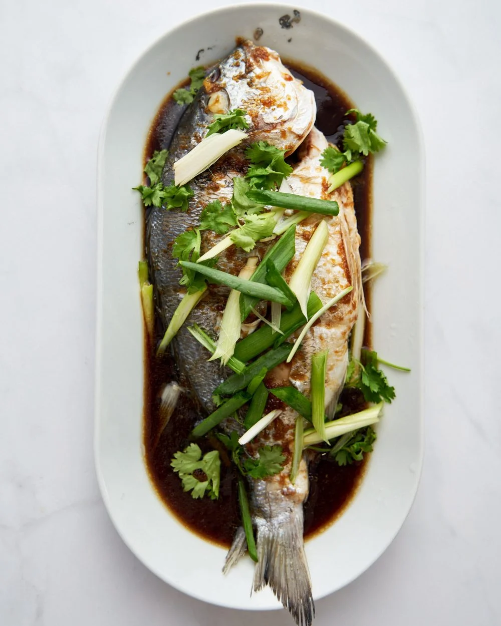 Whole dorado fish in a white oval dish with soy sauce and long cut green onions all over it.