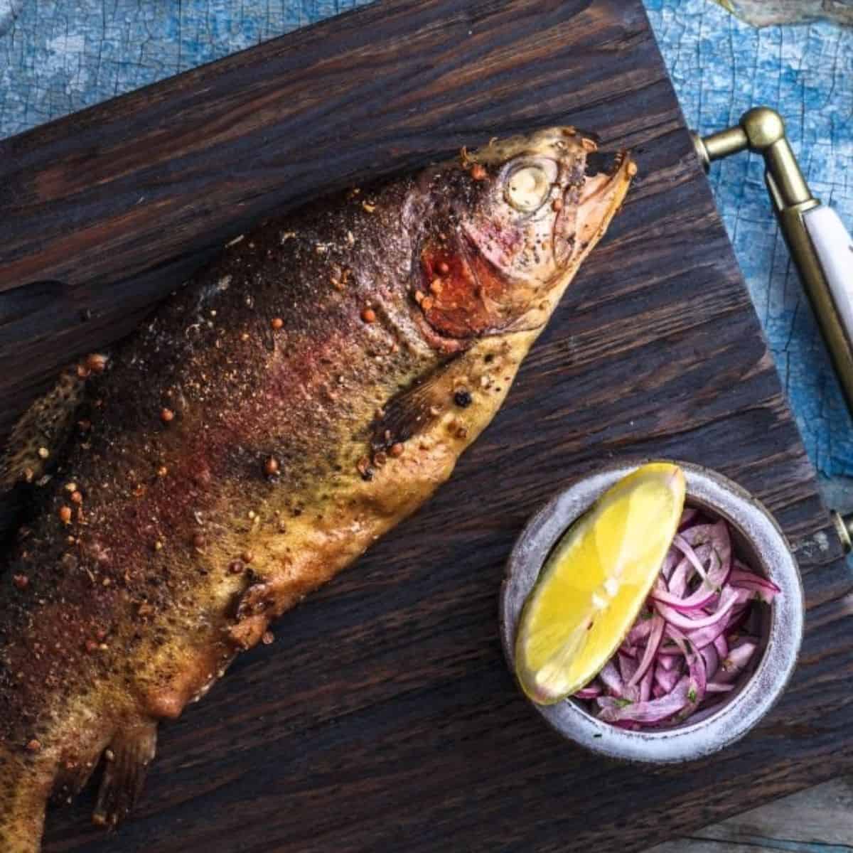 Whole smoked trout on a dark wood board with lemon wedge and red onion beside.