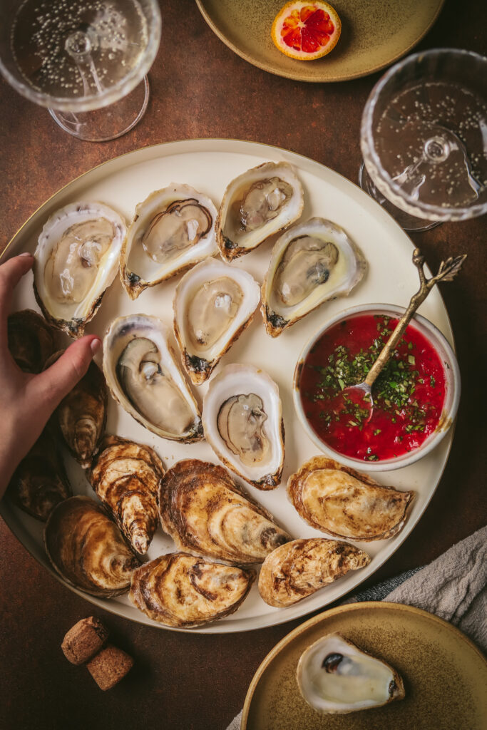 White round platter with raw oysters on the half shell and a bowl of red mignonette sauce beside.