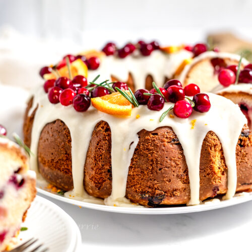 Side view of the white glazed bundt cake topped with red cranberries and triangular orange segments on a white plate.