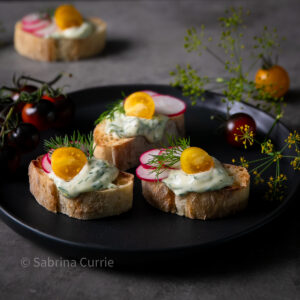Black plate of bruschetta topped with creamy pale green aioli, sliced radishes and halved yellow cherry tomatoes.