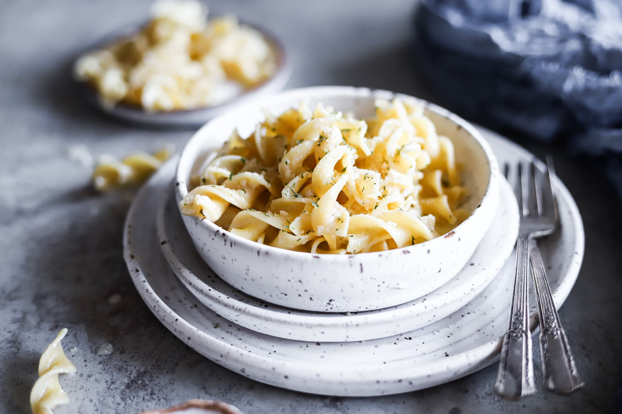 Creamy buttered fettuccini noodles in a white bowl on a dark grey table cloth.