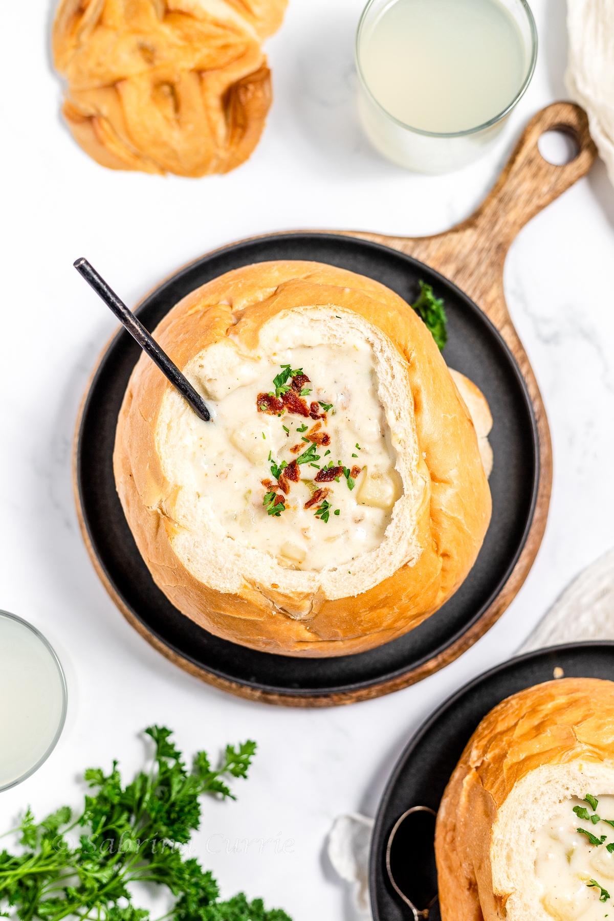 Individual bread bowl hollowed out and filled with clam chowder in the middle on a black plate set on a wooden board. There is a spoon in the soup and a sprinkling of crumbled bacon and chopped parsley on top.