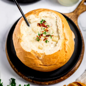 Individual bread bowl hollowed out and filled with clam chowder in the middle. There is a spoon in the soup and a sprinkling of crumbled bacon and chopped parsley on top.