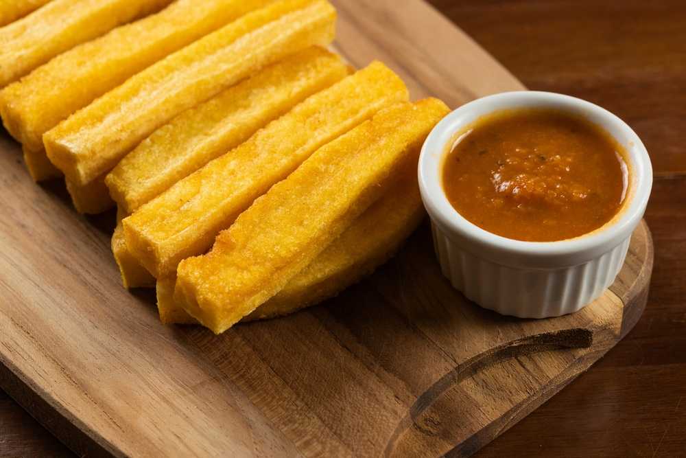 Crispy yellow sticks of polenta on a wooden board with a dark reddish brown dipping sauce beside.