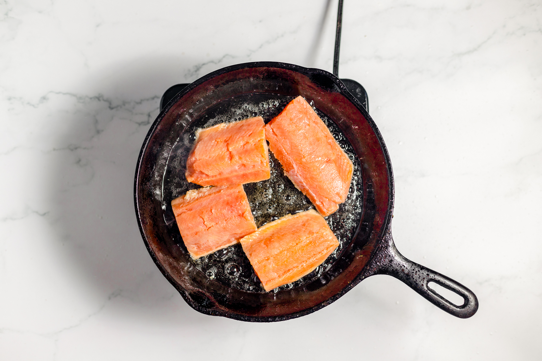 4 large pieces of salmon in a black cast iron frying pan with bubbling oil around it.