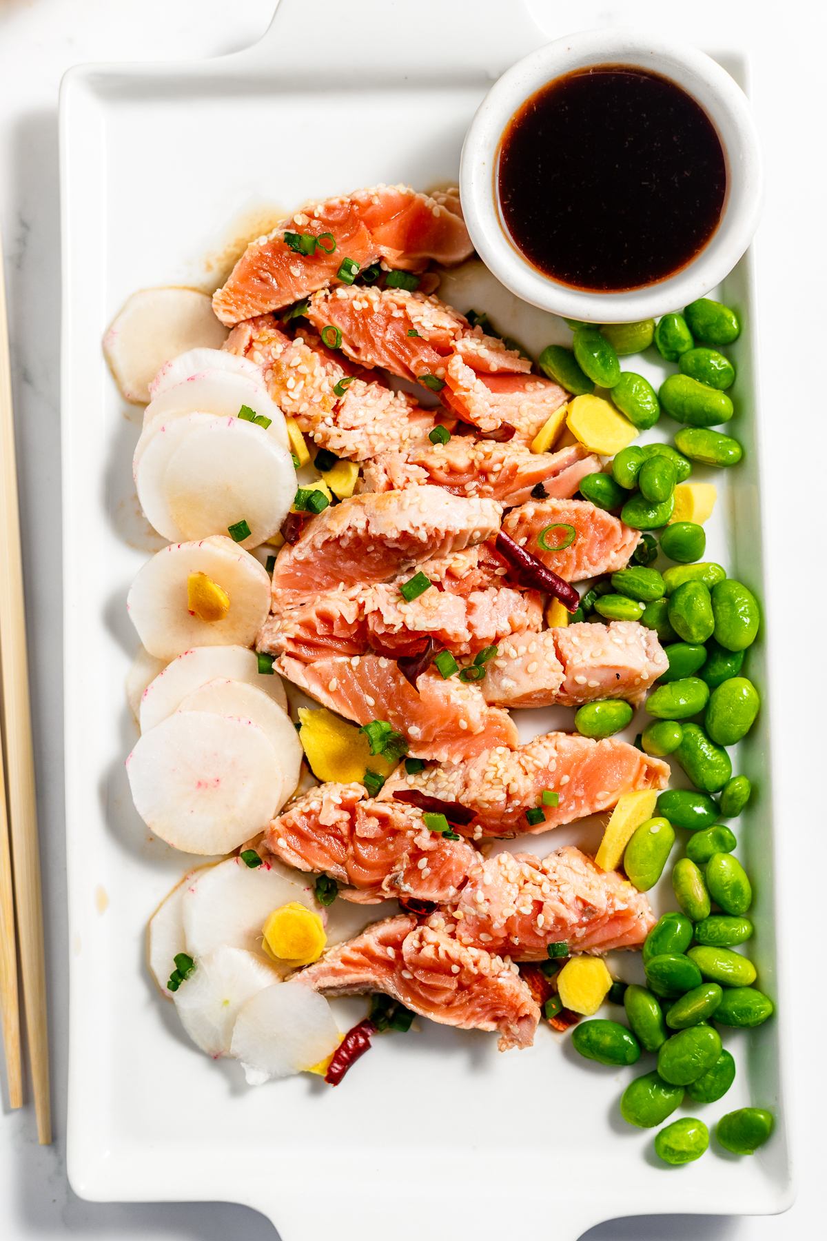 Platter of red salmon tataki pieces in the center flanked by Japanese edamame beans on the right and slivered white daikon on the left. There is a small bowl of dark ponzu sauce in the top right corner.