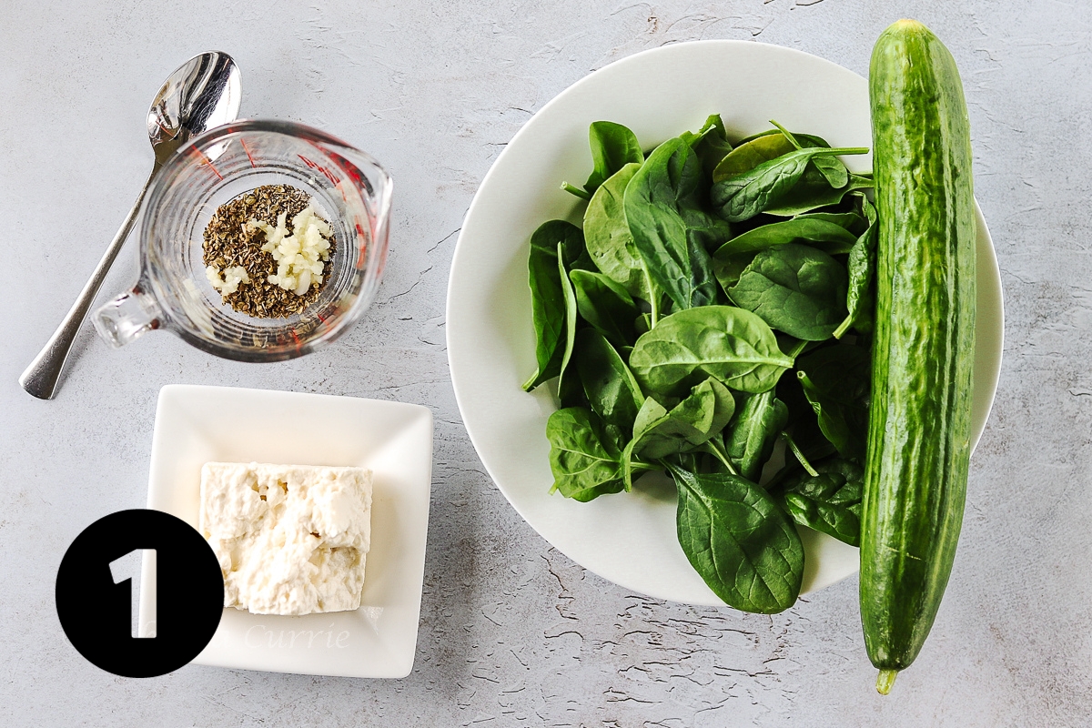 A glass measuring cup with dried herbs and crushed garlic in it, alongside a bowl of spinach and a cucumber and a square dish with some feta cheese.
