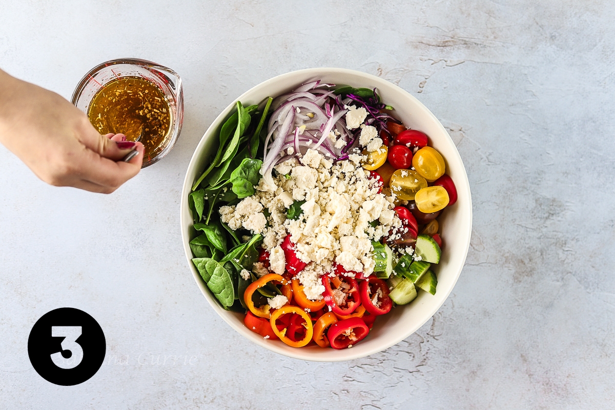 A large salad bowl filled with spinach leaves, chopped tomatoes, cucumber, peppers, and red onion topped with white crumbled feta cheese. There is a hand stirring the homemade salad dressing in the glass measuring cup beside.