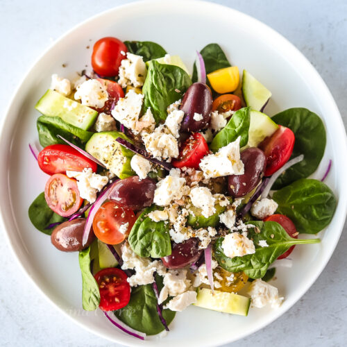 Greek spinach salad with baby spinach leaves, halved cherry tomatoes, chopped cucumbers, kalamata olives, feta cheese and a herb flecked pale yellow dressing drizzled over, all on a white plate.