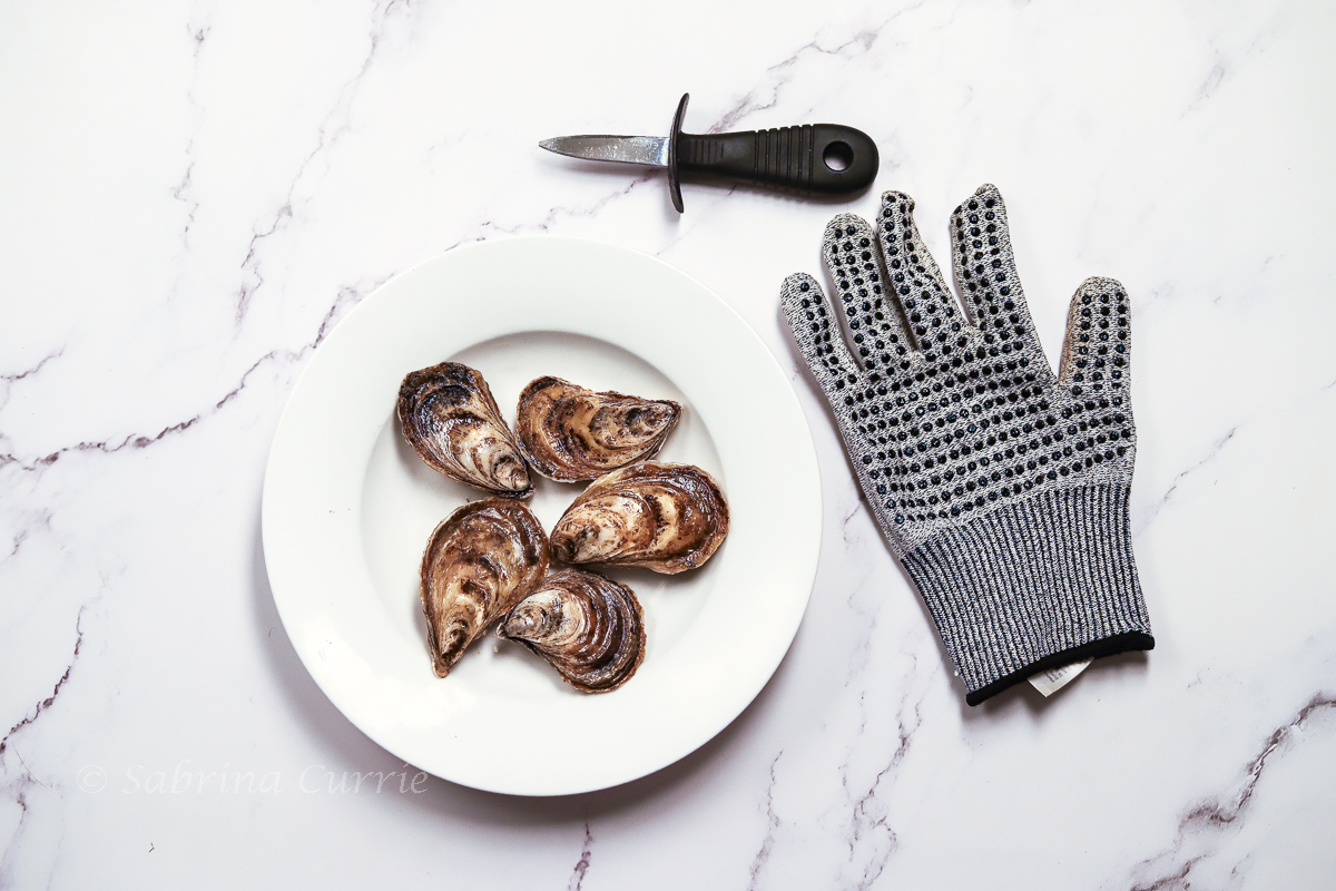 Whole oysters in a bowl with a chain mail oyster shucking glove and oyster knife beside.