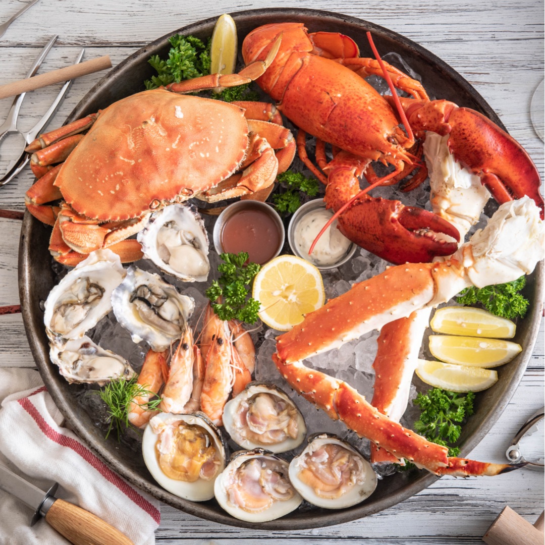 Round black platter of seafood including a crab, a lobster, crab legs, oysters and clams on the half shell and wedges of lemon.