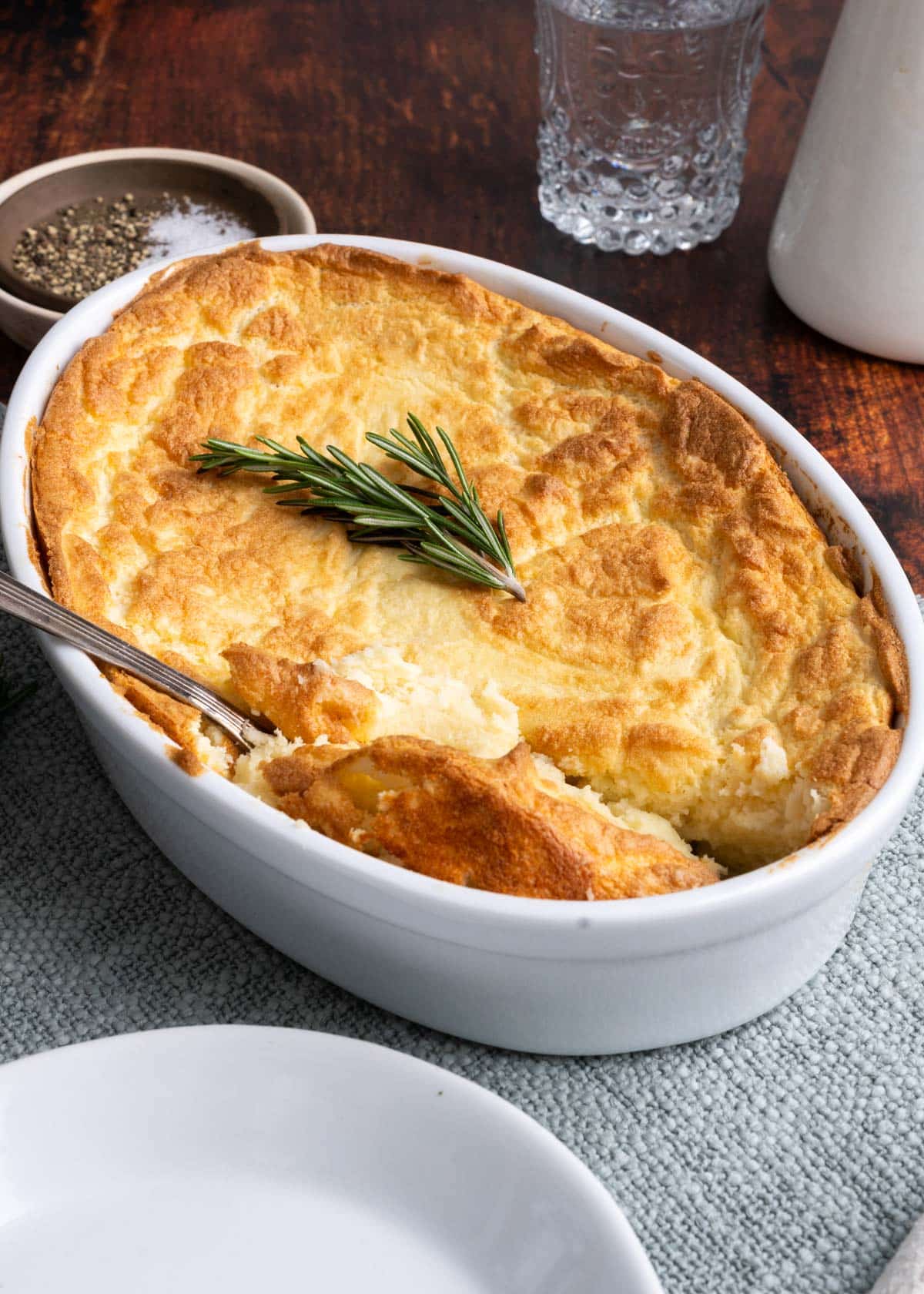 White oval casserole dish with a lightly browned puffy potato souffle topped with a sprig of rosemary.