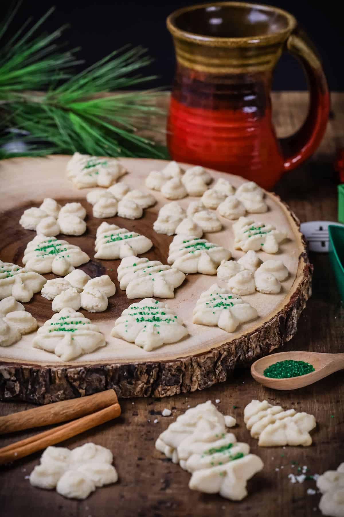 White Christmas tree shaped spritz cookies on a round wooden board. The cookies are topped with some green sugar sprinkles.