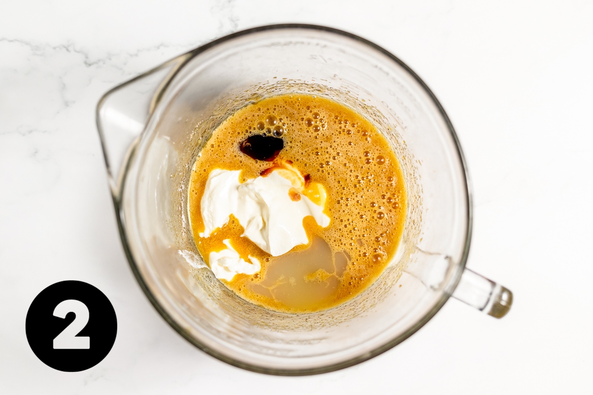 Dark brown vanilla and white sour cream have been add to the yellow egg mixture in the glass measuring cup.