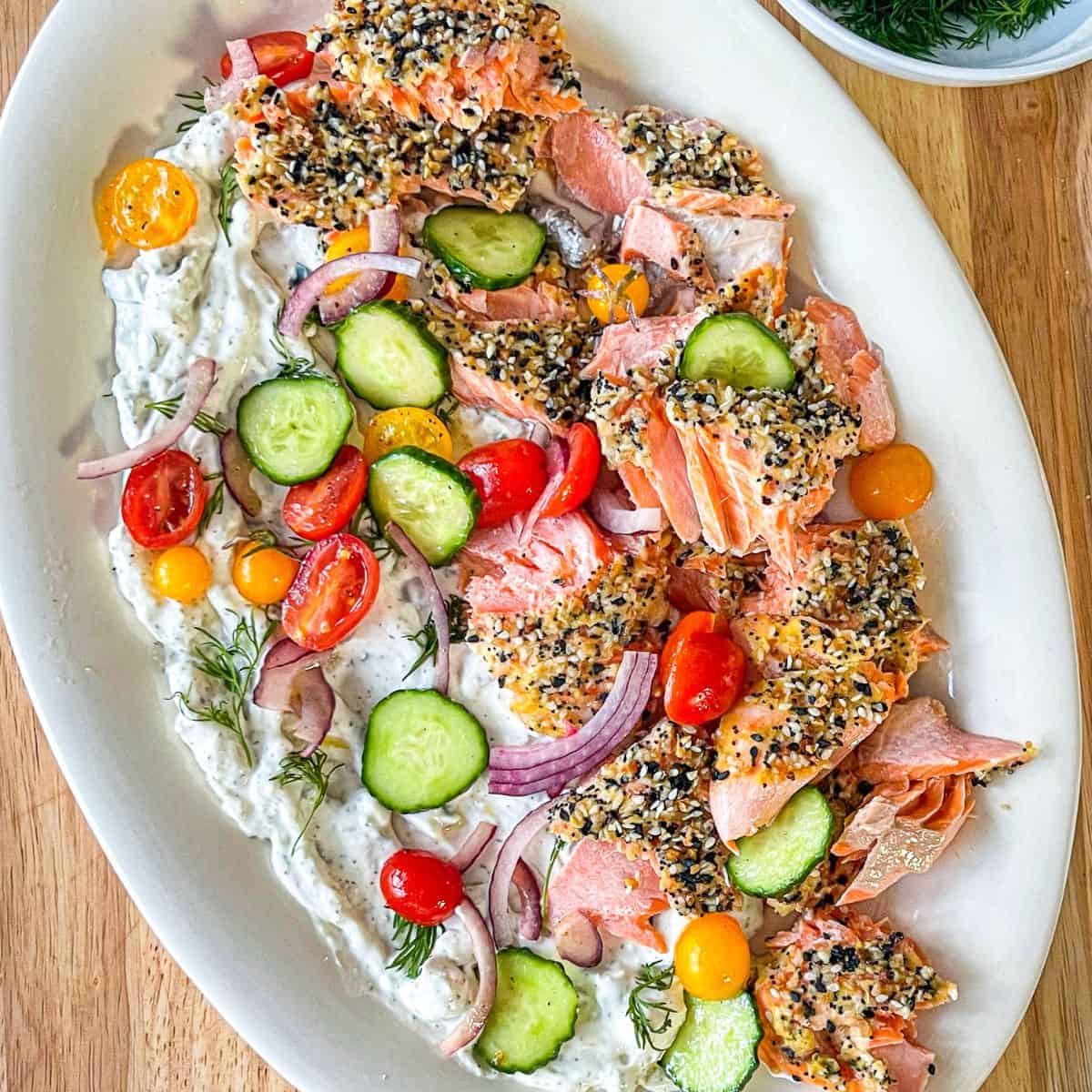 White oval platter with pale pink baked steelhead trout, creamy green flecked tsatsiki sauce and chopped tomatoes and cucumbers.